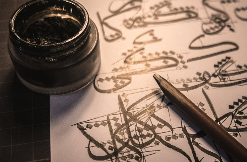 A traditional dipping wooden pen or qalam, with a bottle of black ink, on a paper with practice sketches of khat or Arabic calligraphy Thuluth script.