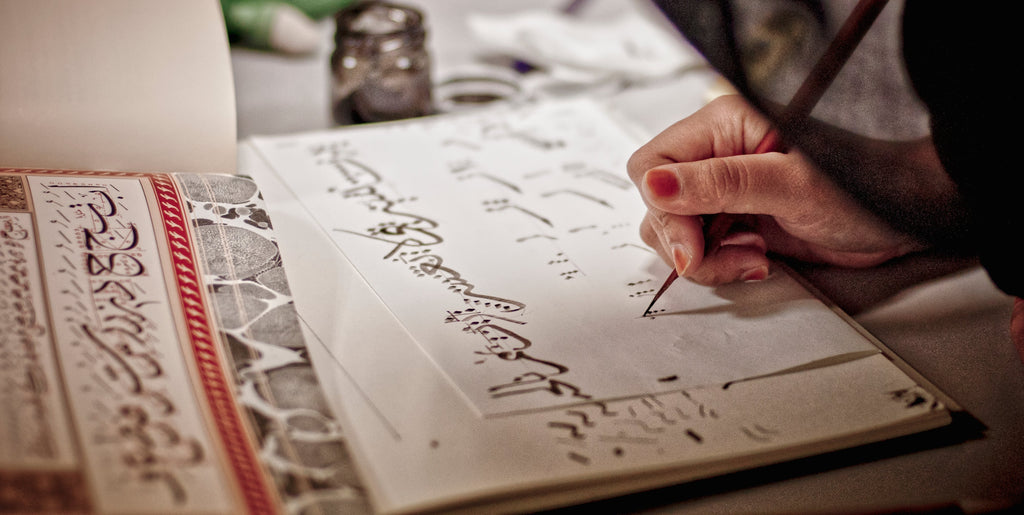 A female student practising traditional khat or Arabic calligraphy by using a wooden pen or qalam to copy a master’s work.