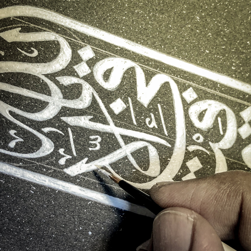 Hand holding a fine brush with white ink painting Islamic calligraphy artwork.